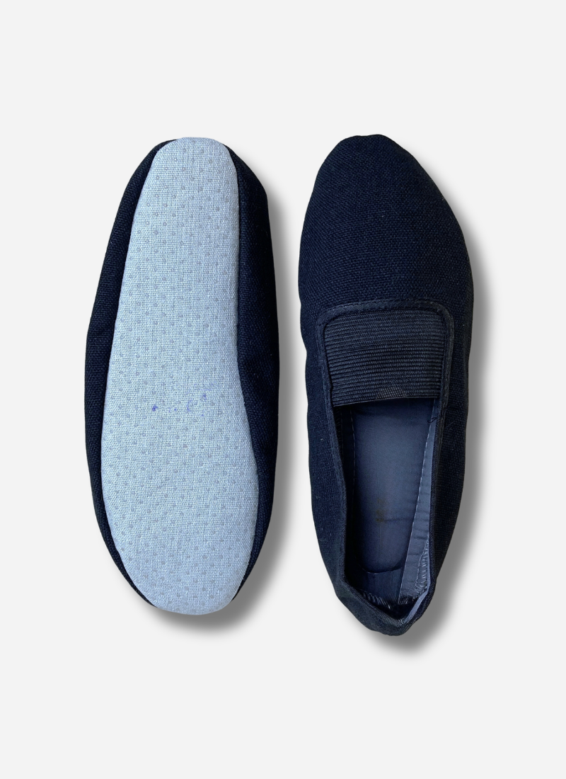 Two black adults canvas gymnastics and dance slippers shown side-by-side against a plain background. One shoe shows the top view with the elastic strap; the other shows the light blue, anti-slip textured sole.