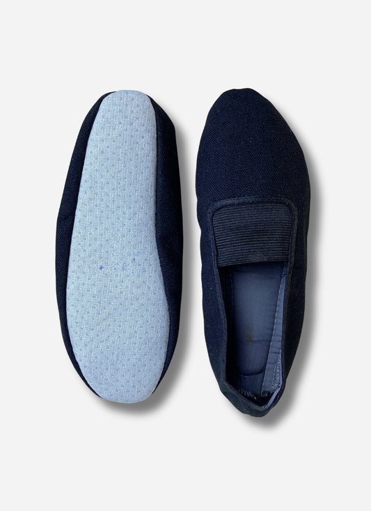 Two black adults canvas gymnastics and dance slippers shown side-by-side against a plain background. One shoe shows the top view with the elastic strap; the other shows the light blue, anti-slip textured sole.