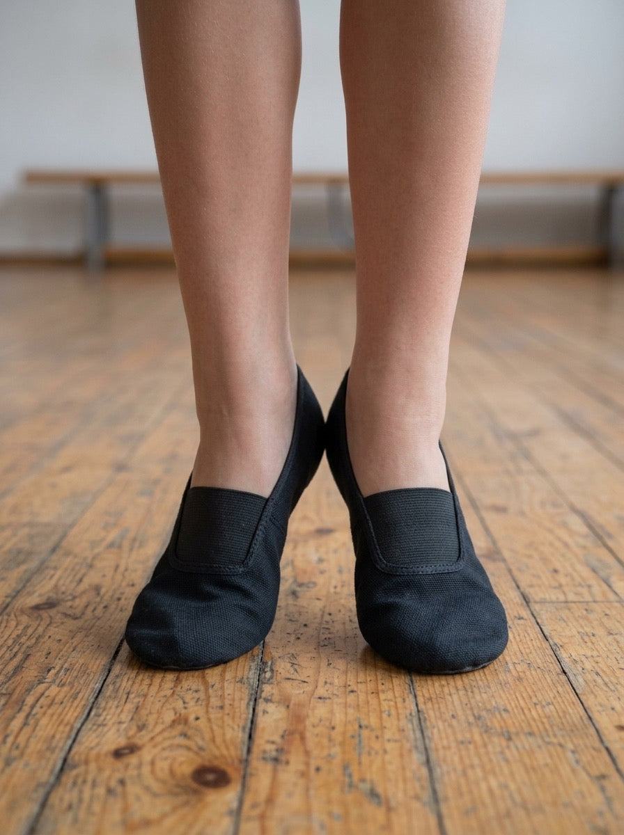 A model's lower legs and feet on a wooden floor, wearing a pair of black canvas gymnastics and dance slippers. A close-up front-on view.