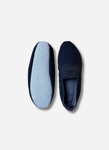 Two black kids canvas gymnastics and dance slippers shown side-by-side against a plain background. One shoe shows the top view with the elastic strap; the other shows the light blue, anti-slip textured sole.