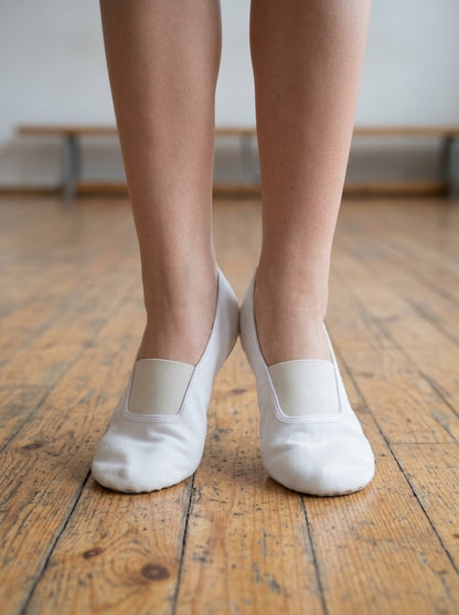 A model's lower legs and feet on a wooden floor, wearing a pair of white canvas gymnastics and dance slippers. A close-up front-on view.