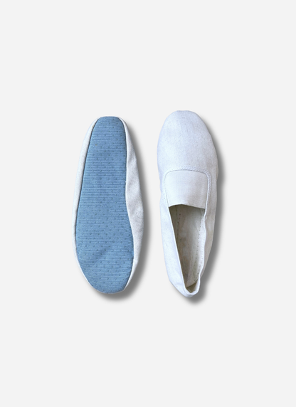 Two white kids canvas gymnastics and dance slippers shown side-by-side against a plain background. One shoe shows the top view with the elastic strap; the other shows the light blue, anti-slip textured sole.