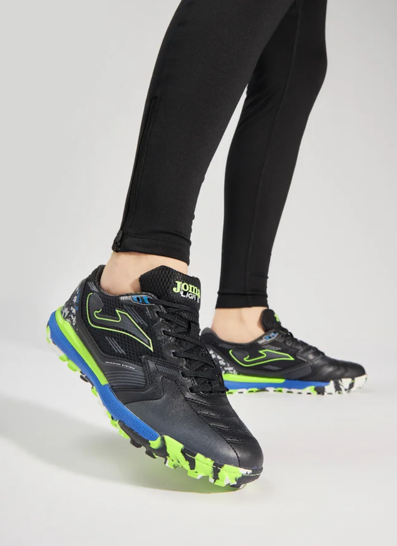 Image 1 (on-feet lifestyle shot) Joma Liga 5 men's turf football boots worn on feet, showing side profile with black upper, royal blue midsole, and lime green accents