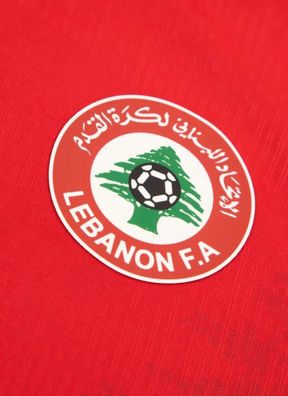 Close-up view of the circular Lebanon Football Association crest featuring a green cedar tree and football on a red textured kit.