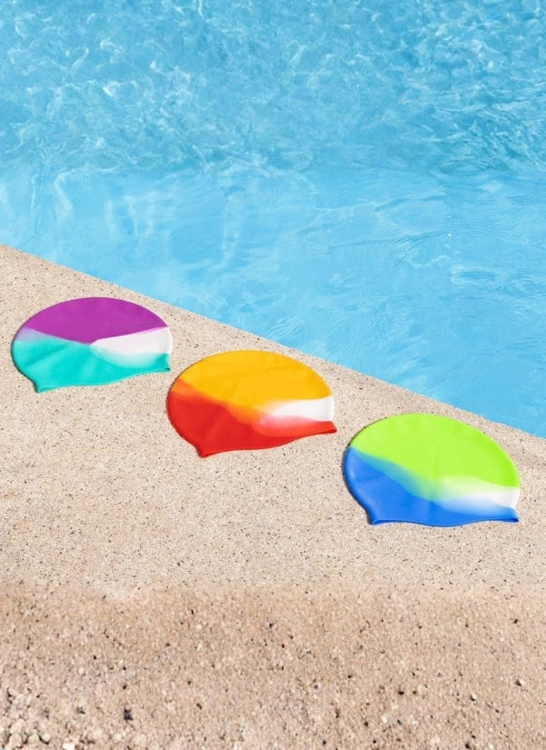 Three colorful swim caps with a rainbow gradient design are lined up on a sandy poolside deck with blue water in the background.