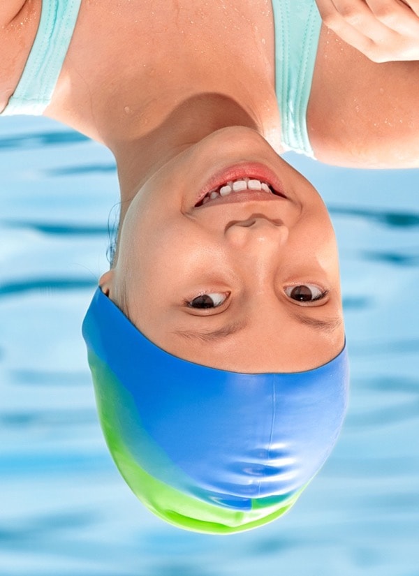 A smiling young girl wearing a blue and green gradient swim cap, with water ripples in the background.