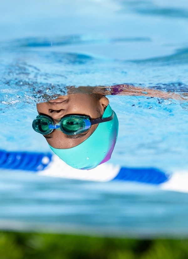 A child swimming in a pool, wearing a bright turquoise swim cap, blue swim goggles, and a purple swimsuit.