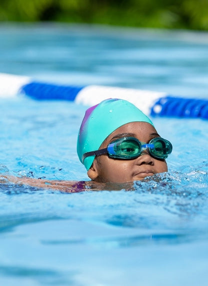A child swimming in a pool, wearing a bright turquoise swim cap, blue swim goggles, and a purple swimsuit.