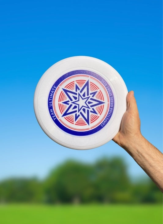 Hand holding up a white flying disc with a blue and red star graphic against a blue sky and green field